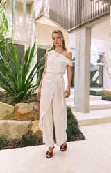 Model wearing a cream House of Holabox one-shoulder draped jumpsuit with a waist tie in a modern outdoor setting.