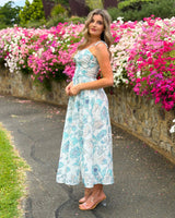House of Holabox Belle maxi dress with blue floral print, twist-front bodice, and tie straps in a garden setting