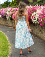 House of Holabox Belle Maxi Dress featuring a blue floral print, tie straps, and a flowing skirt in a garden setting.