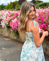 House of Holabox Belle maxi dress with blue floral print, tie straps, and smocked back, worn by a woman in a garden.