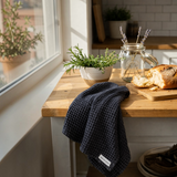 House of Holabox charcoal organic cotton waffle tea towel draped on a rustic wooden kitchen counter by a window.
