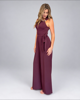 House of Holabox cherry chocolate jumpsuit with asymmetric neckline, gold buckle detail, and waist tie on a model.