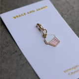 Cupcake shaped gold-plated enamel hanging charm with pink and white painted details on a branded display card.