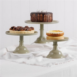 House of Holabox Ecology Torte glass cake stands in sage green with various cakes displayed on a white tablecloth.