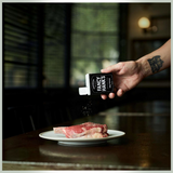 House of Holabox Fancy Hank's BBQ Pepper seasoning being sprinkled from a 90g tin onto a raw steak on a plate