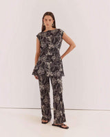House of Holabox Fiora tunic top in black with white floral print, boat neckline, and waist tie, worn with trousers.