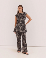 House of Holabox Fiora tunic top in black with white floral print, boat neckline, and waist tie, worn with trousers.