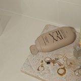 House of Holabox Ivy Ila Japanese Honeysuckle body custard in a beige bottle on a stone surface with gold jewelry.
