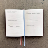 House of Holabox Jour Studio linen planner open to a monthly reflection page with blue and terracotta ribbon markers.