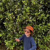 A smiling child wearing House of Holabox Little Drop round orange sunglasses and a matching cap outdoors.