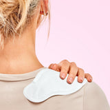 A person applying a white House of Holabox Lula self-warming body patch to their shoulder over a beige top.