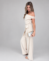 House of Holabox one-shoulder cream jumpsuit with draped waist tie worn by a model holding a patterned clutch