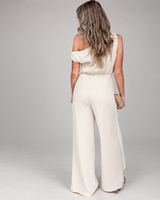 House of Holabox one-shoulder cream jumpsuit with draped waist detail, worn by a woman with long wavy hair.