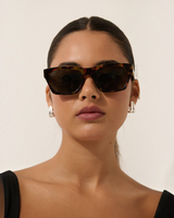 House of Holabox Ria square sunglasses in tort acrylic with dark tinted lenses worn by a woman with silver earrings.