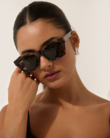 House of Holabox Ria square sunglasses in tort acrylic with dark tinted lenses worn by a woman against a white wall.