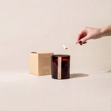 House of Holabox saide. large amber soy wax candle with a wood wick being lit, beside its signature earthy-toned box.
