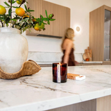 House of Holabox saide. amber glass wood wick candle burning on a marble kitchen counter with a blurred woman behind.