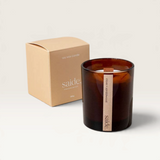 House of Holabox saide. large amber glass soy candle with a wood wick next to its matching earthy-toned gift box.