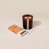 House of Holabox saide. large amber glass soy candle with a wood wick and a matching box of matches on a beige surface.