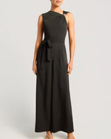 A woman modeling a sleeveless black one-shoulder maxi dress with a draped neckline, a matching fabric tie belt at the waist, and wide legs, photographed against a light gray studio background.