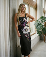 Woman in a floral dress standing in a room with large windows and plants.