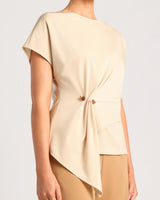 A woman wearing a light beige short-sleeve top with a twisted front detail and tan pants, photographed against a plain light gray studio background.
