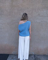 Woman wearing a blue asymmetrical moss knit top paired with white wide-leg trousers against a textured wall.