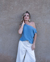 Woman wearing a blue asymmetrical moss knit top paired with white wide-leg trousers against a textured wall.