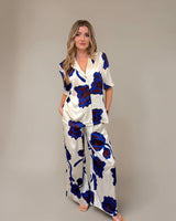 Woman wearing a cream satin camp collar shirt and matching wide-leg trousers with a bold blue and dark floral print.