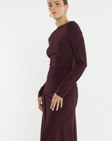 Model wearing a long-sleeve dark burgundy jersey dress with a boat neckline, gathered waist, and seam details.