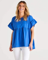 Blue Jameson cotton blouse with a scoop neck, empire waist, and mini frill sleeves worn with white pants.