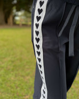 Black Jamie Jogger trackpants featuring a white side stripe with a repeating black heart pattern and a drawstring waist.