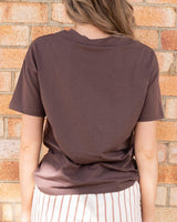 Back view of a chocolate brown cotton-blend crew neck t-shirt worn against a rustic brick wall.