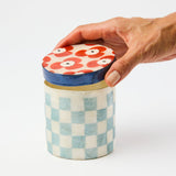 Hand holding a checkered container with a floral lid on a white background