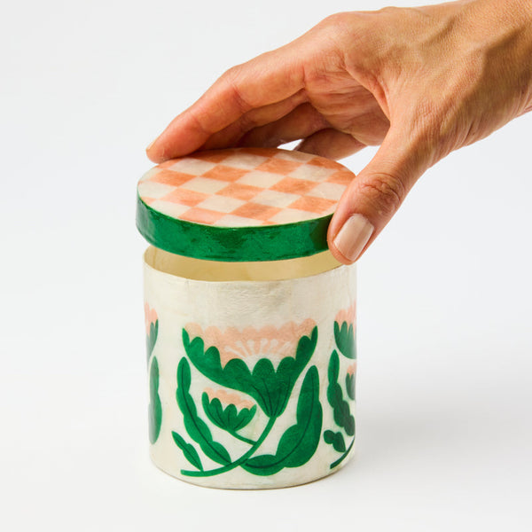 Hand holding a decorative ceramic jar with a green lid on a white background