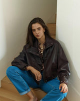 Woman wearing a dark brown leather-look bomber jacket with a high neck, zip front, and relaxed fit on wooden stairs.