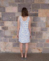 Woman wearing a blue and white embroidered shift dress with wide straps, standing against a stone wall.