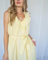 Woman wearing a pale yellow cotton sun dress with a ruffled neckline and a self-tie waist belt.