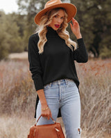 Woman wearing a black high-neck knit sweater with ribbed cuffs and a high-low hem, paired with light wash distressed jeans.