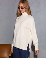 Woman wearing a cream high-neck knit sweater with long sleeves and side slits, styled with blue jeans and sunglasses.