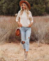 Woman wearing a cream high-neck knit sweater with long sleeves, styled with distressed jeans and a brown fedora hat.
