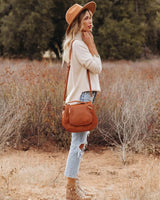 Woman wearing a cream high-neck knit sweater with a brown hat, crossbody bag, and jeans in a field.