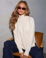 Woman wearing a cream high-neck knit sweater with long sleeves and ribbed trim, styled with dark blue jeans.