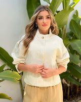 Woman wearing a cream-colored cotton Lori jacket with a high collar and button-front closure against green foliage.