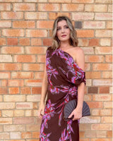 Woman wearing a brown one-shoulder maxi dress with a purple and pink floral print and gathered waist detail.