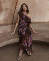Model wearing a brown one-shoulder maxi dress in viscose yoryu with a purple and red floral print and waist gathering.