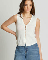 Woman wearing a white, sleeveless, V-neck knit vest with a front hook-and-eye closure paired with blue jeans.