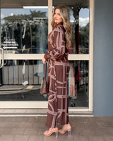 Woman wearing a brown long-sleeve maxi dress with a white geometric print, styled with clear heels and a woven bag.