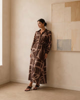 Model wearing a brown long-sleeve maxi dress with a geometric cream print, button-down front, and tie waist.