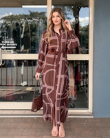 Woman wearing a brown long-sleeve maxi dress with a white geometric print, button-down front, and tie waist.